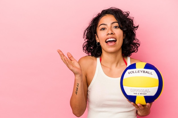 Молодая латинская женщина, держащая волейбольный мяч на розовом фоне, показывает пространство для копии на ладони и держит другую руку на талии.