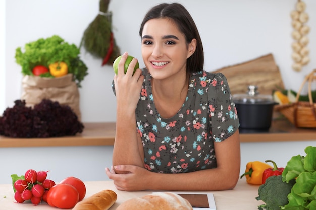 젊은 라틴 여성이 녹색 사과를 들고 태블릿 컴퓨터로 온라인 쇼핑을 하고 있습니다. 주부는 부엌에서 새로운 요리법을 발견했습니다