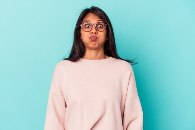Young latin woman isolated on blue background blows cheeks, has tired expression. Facial expression concept.