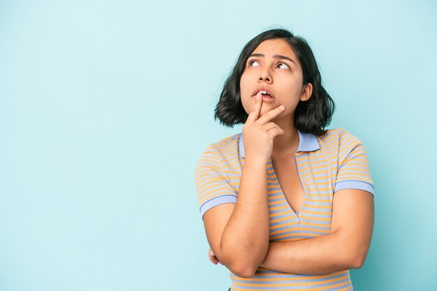 Молодая латинская женщина, изолированная на синем фоне, смотрит в сторону с сомнительным и скептическим выражением лица.