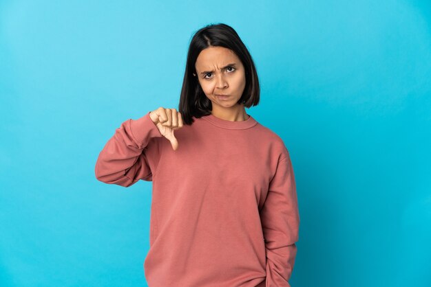Молодая латинская женщина изолирована на синей стене, показывая большой палец вниз с отрицательным выражением лица