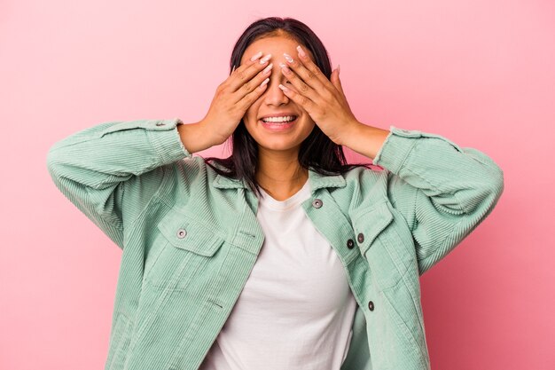 La giovane donna latina isolata su sfondo rosa copre gli occhi con le mani, sorride ampiamente in attesa di una sorpresa.