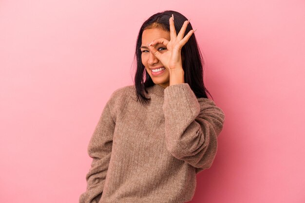 Молодая латинская женщина, изолированная на розовом фоне, взволнована, держа жест ок на глазу