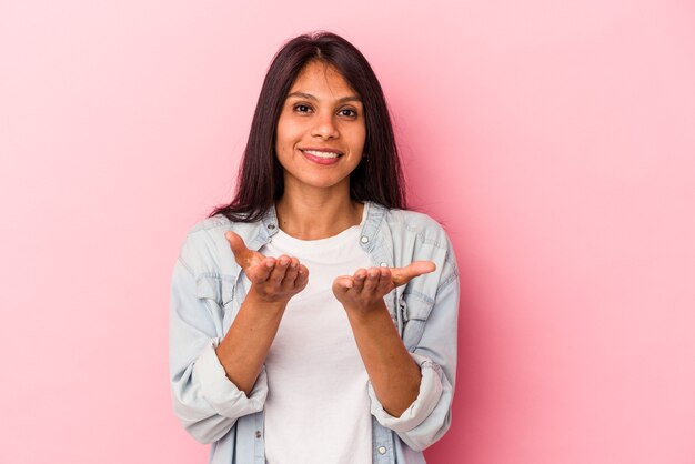 Giovane donna latina isolata su sfondo rosa che tiene qualcosa con le palme, offrendo alla telecamera.