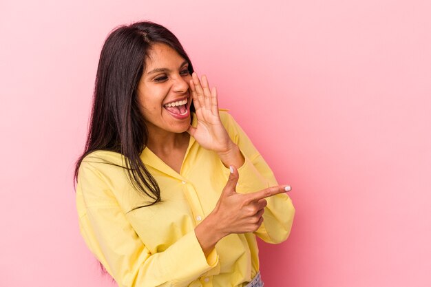 Young latin woman isolated on pink background saying a gossip, pointing to side reporting something.