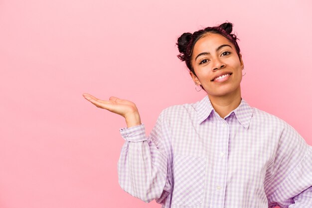 Young latin woman isolated on pink background showing a copy space on a palm and holding another hand on waist.