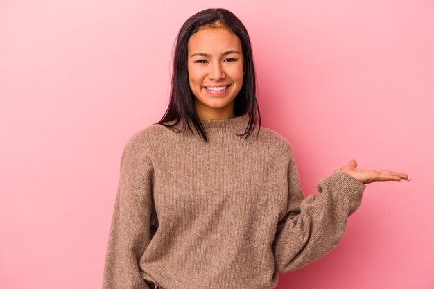 Young latin woman isolated on pink background  showing a copy space on a palm and holding another hand on waist.