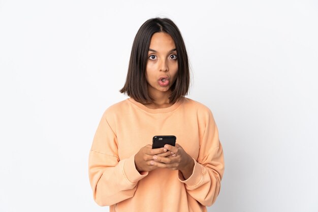 Young latin woman isolated on white wall looking at the camera while using the mobile with surprised expression