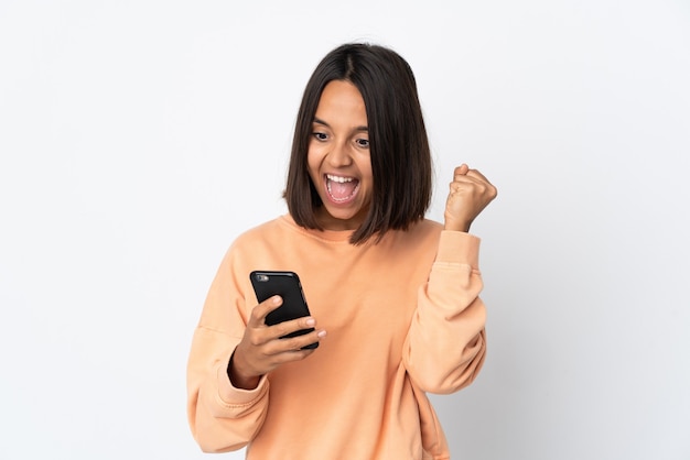 Young latin woman isolated on white wall using mobile phone and doing victory gesture