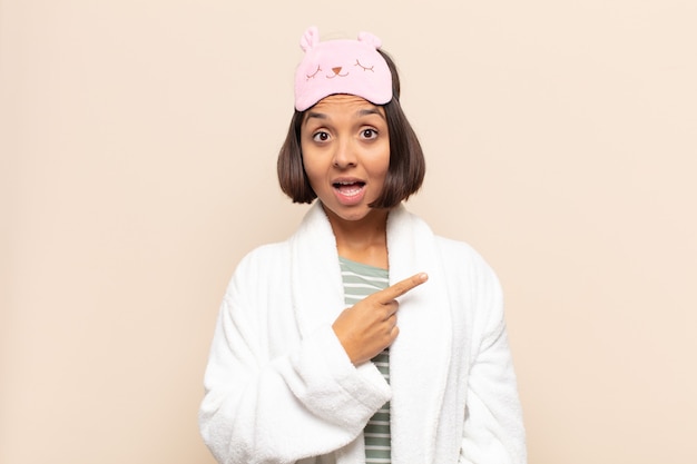 Young latin woman looking excited and surprised pointing to the side and upwards to copy space