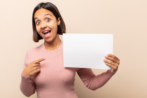 Young latin woman looking excited and surprised pointing to the side and upwards to copy space