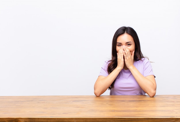 Young latin woman looking happy, cheerful, lucky and surprised covering mouth with both hands on flat wall