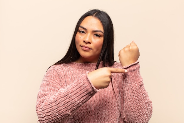 Young latin woman looking impatient and angry, pointing at watch, asking for punctuality, wants to be on time