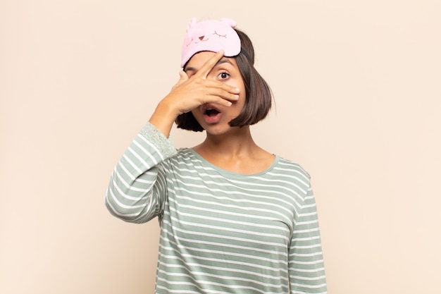 Young latin woman looking shocked, scared or terrified, covering face with hand and peeking between fingers