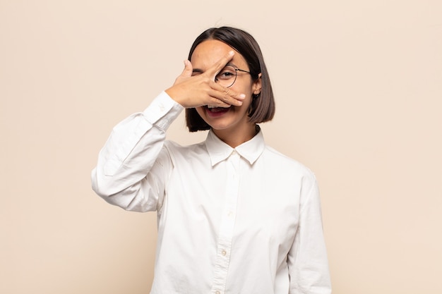 Young latin woman looking shocked, scared or terrified, covering face with hand and peeking between fingers