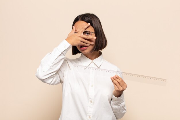 Young latin woman looking shocked, scared or terrified, covering face with hand and peeking between fingers