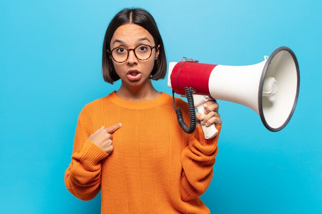 Young latin woman looking shocked and surprised with mouth wide open, pointing to self