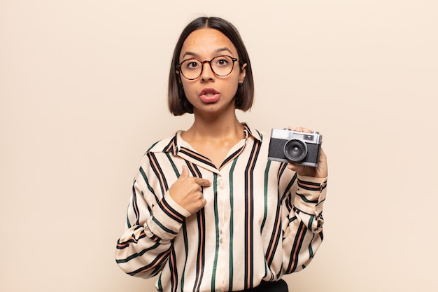 young latin woman looking shocked and surprised with mouth wide open, pointing to self