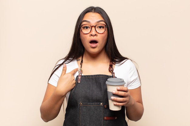 Young latin woman looking shocked and surprised with mouth wide open, pointing to self