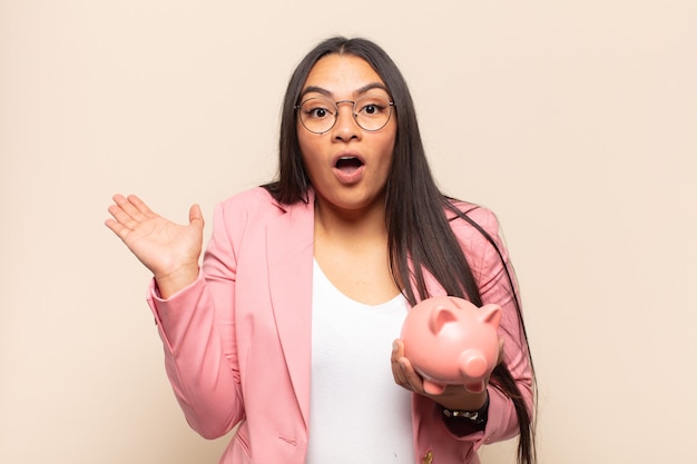 Young latin woman looking surprised and shocked, holding a piggy bank