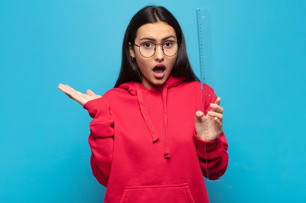 Young latin woman looking surprised and shocked, with jaw dropped holding an object with an open hand on the side