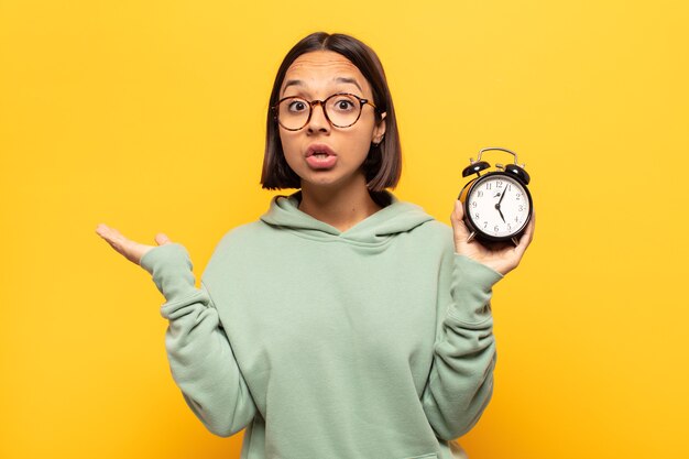 Young latin woman looking surprised and shocked, with jaw dropped holding an object with an open hand on the side