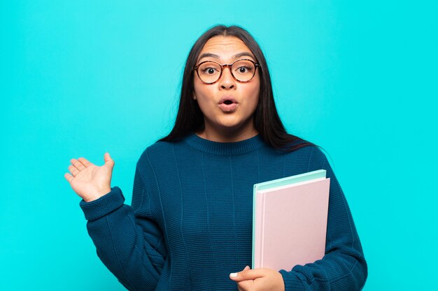 Young latin woman looking surprised and shocked, with jaw dropped holding an object with an open hand on the side