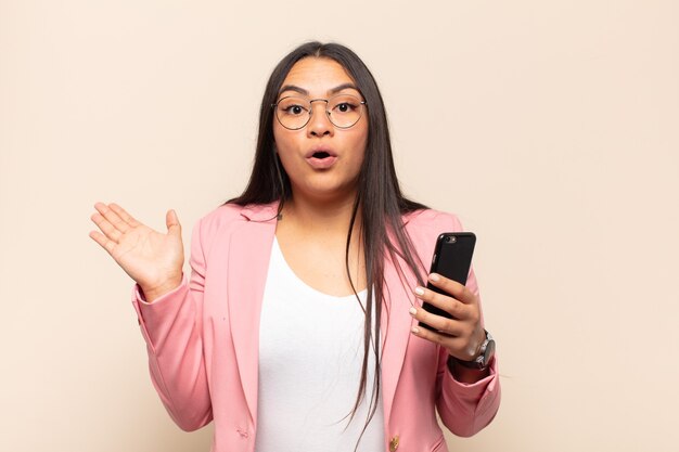 Young latin woman looking surprised and shocked, with jaw dropped holding an object with an open hand on the side