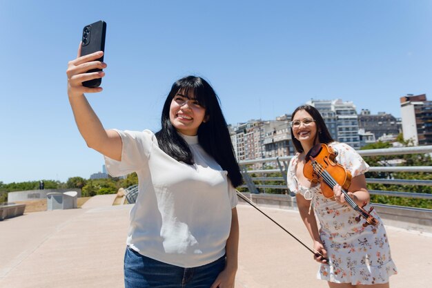 young latin woman outdoors happy taking selfie photo with street violinist artist