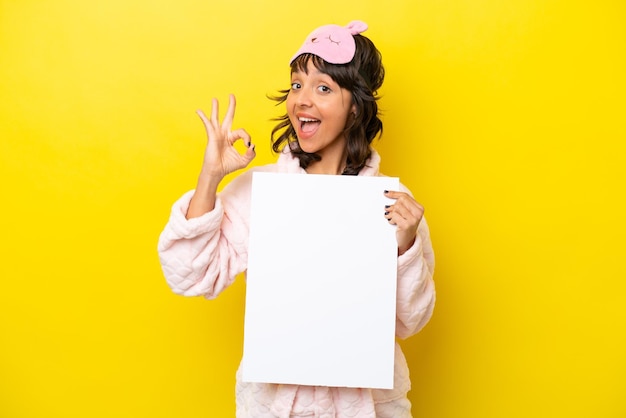 Молодая латиноамериканка в пижаме на желтом фоне держит пустой плакат и делает знак ОК
