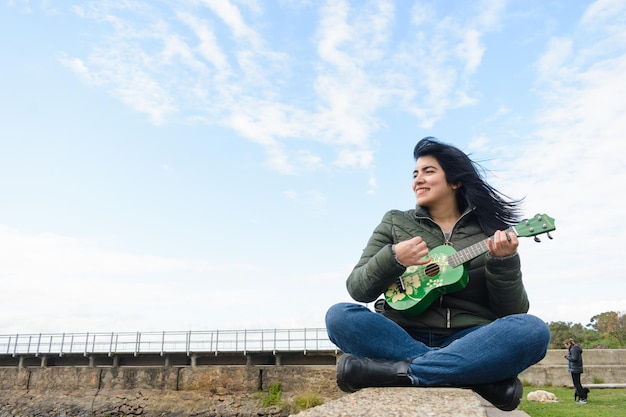 Молодая латиноамериканка сидит на открытом воздухе и играет на укулеле, наслаждаясь прогулкой в выходные утра.