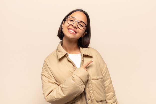 Young latin woman smiling cheerfully, feeling happy and pointing to the side and upwards, showing object in copy space