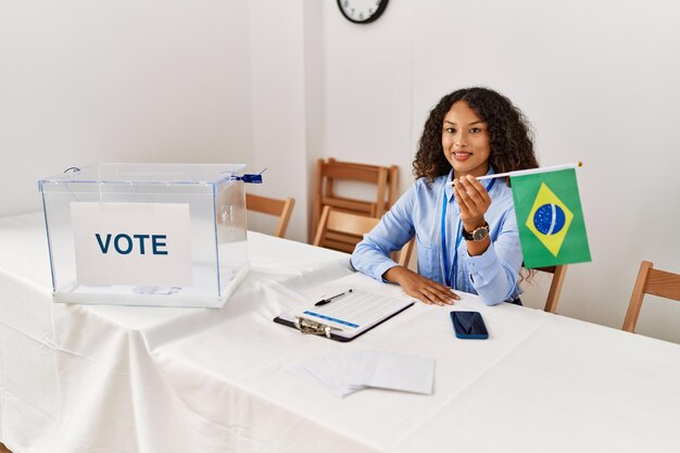 Young latin woman smiling confident holding brazil flag working at electoral college