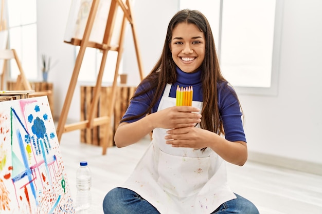 Photo young latin woman smiling confident holding color pencils at art studio