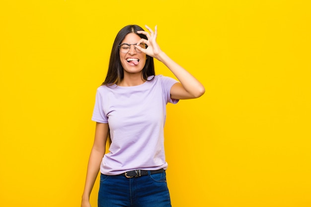 Young latin woman smiling happily with funny face, joking and looking through peephole, spying on secrets on flat wall