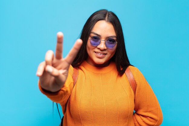 Young latin woman smiling and looking happy, carefree and positive, gesturing victory or peace with one hand