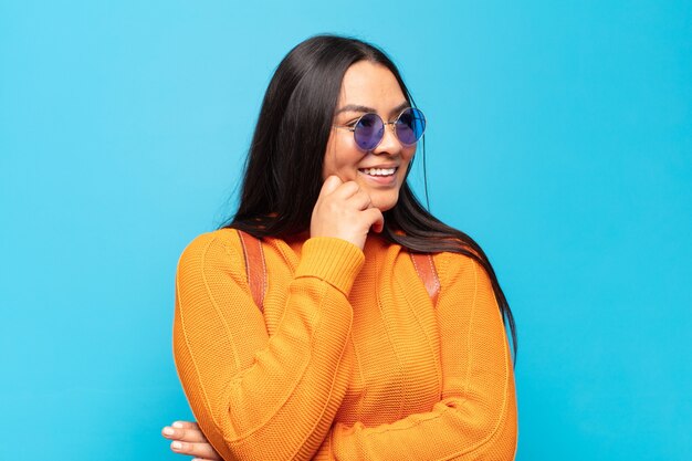 Young latin woman smiling with a happy, confident expression with hand on chin, wondering and looking to the side