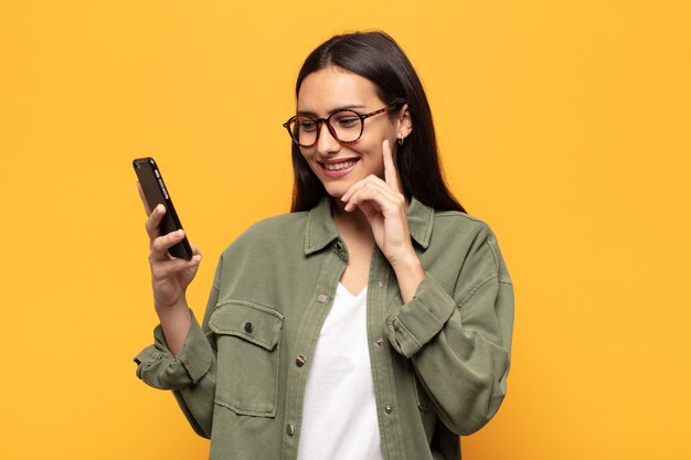 Young latin woman smiling with a happy, confident expression with hand on chin, wondering and looking to the side