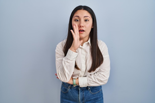 Young latin woman standing over blue background hand on mouth telling secret rumor whispering malicious talk conversation