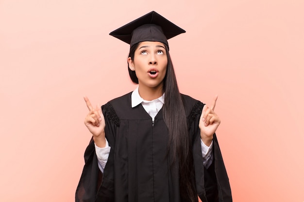 Young latin woman student feeling surprised and open mouthed pointing upwards