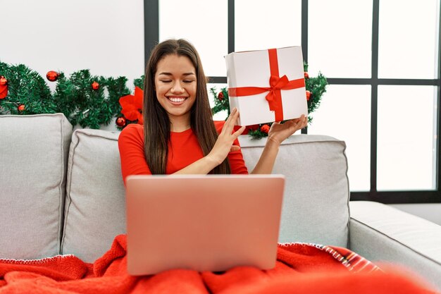 Young latin woman using laptop and holding gift sitting by christmas decor at home