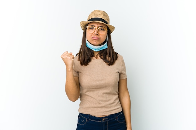 Молодая латинская женщина в шляпе и маске для защиты от covid изолирована на белой стене, показывая кулак агрессивное выражение лица.