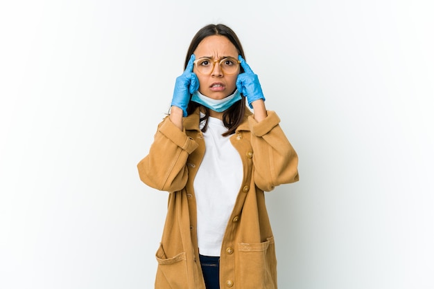 Young latin woman wearing a mask to protect from covid isolated on white wall focused on a task, keeping forefingers pointing head.