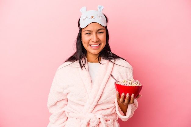Giovane donna latina che indossa un pigiama in possesso di una ciotola di cereali isolata su sfondo rosa felice, sorridente e allegra.