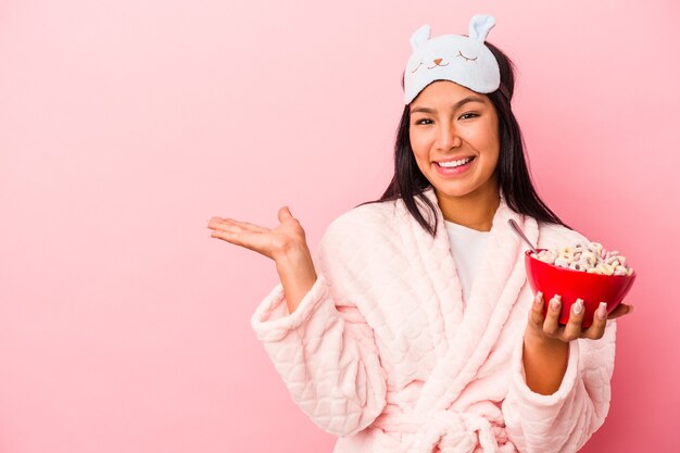 Giovane donna latina che indossa un pigiama che tiene una ciotola di cereali isolata su sfondo rosa che mostra uno spazio di copia su un palmo e tiene un'altra mano sulla vita.