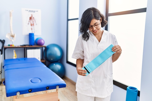 Foto giovane donna latina che indossa l'uniforme del fisioterapista che tiene l'elastico presso la clinica di fisioterapia