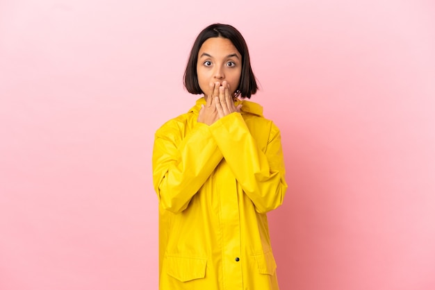 Young latin woman wearing a rainproof coat over isolated background covering mouth with hand