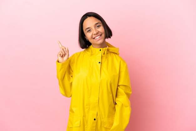 Young latin woman wearing a rainproof coat over isolated background happy and pointing up