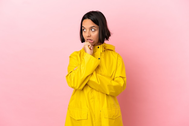 Young latin woman wearing a rainproof coat over isolated background having doubts while looking up