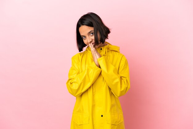 Young latin woman wearing a rainproof coat over isolated background keeps palm together. Person asks for something